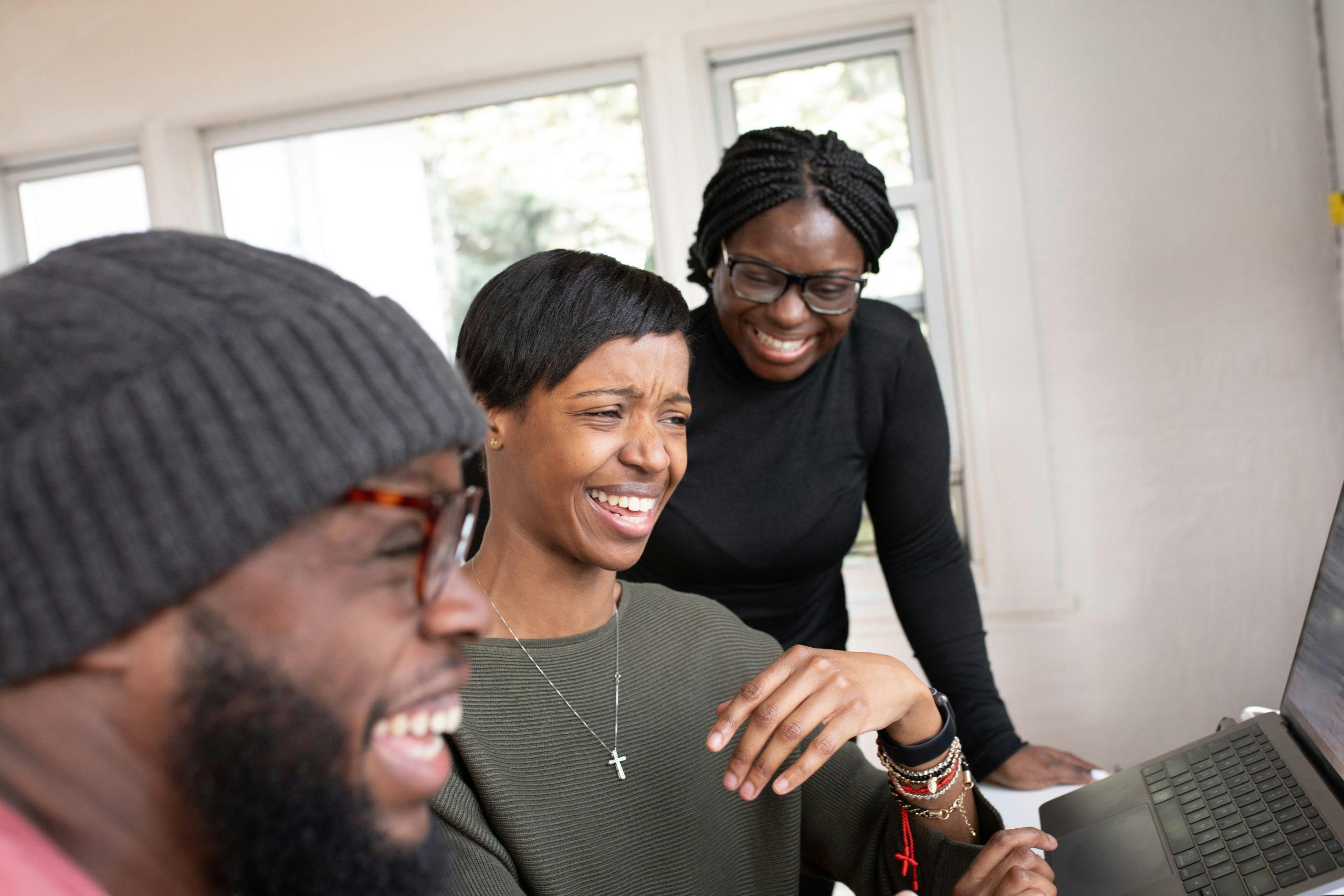 Three friends share a joyful moment while interacting with a laptop indoors.